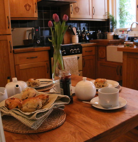 Scones with jam, cream, cups, vase of tulips and teapot on table in kitchen